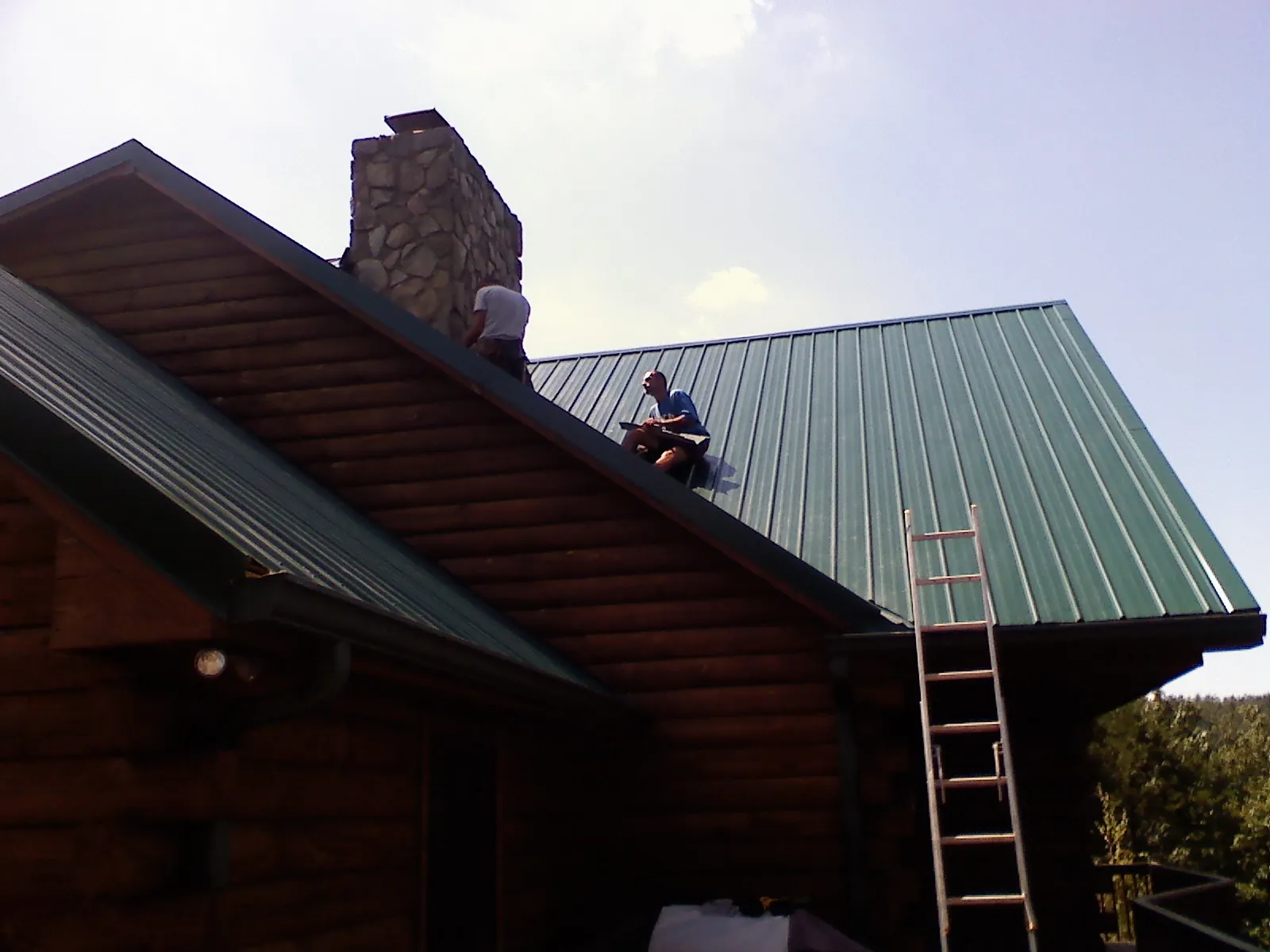 Roofers installing green metal panels beside a stone chimney on a log home in Hingham