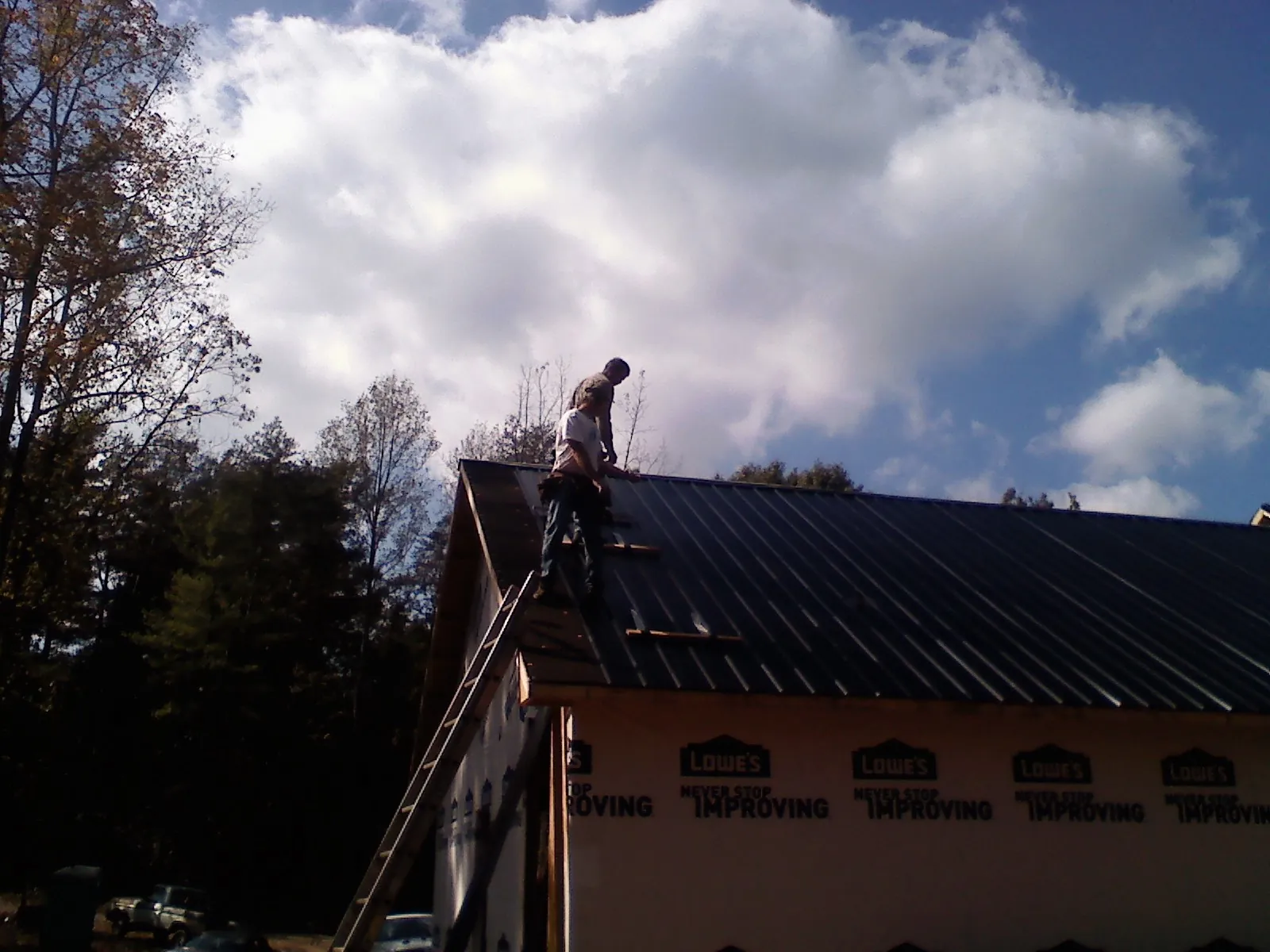 Roofer fastening dark metal panels high against a cloudy sky, from a Roof Inspection job in Hingham